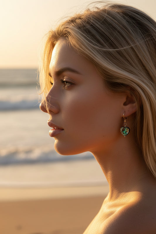 A close-up lifestyle shot of a model's ear wearing a small heart-shaped dangling earring with a colourful, abalone shell heart beads and silver hooks.