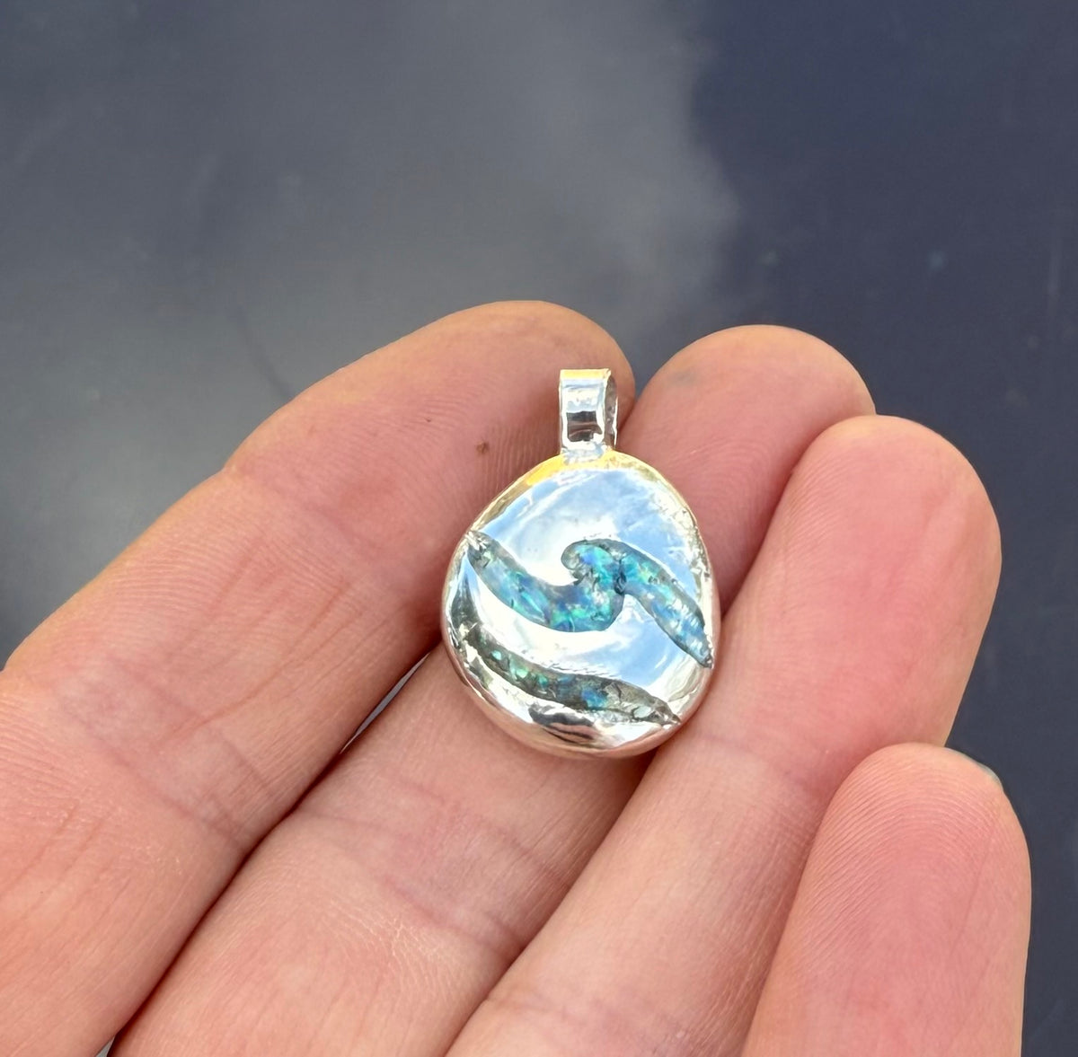 A silver pendant with a blue and white wave design, held in a person's hand against a dark background.