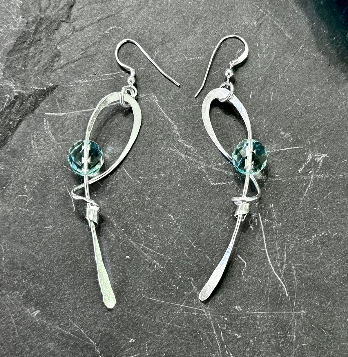 A pair of silver dangle earrings with curved openwork metal loops, a faceted light blue-green bead, and a dangling silver blade, displayed on a dark textured slate surface.