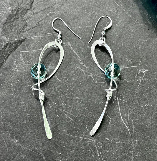 A pair of silver dangle earrings with curved openwork metal loops, a faceted light blue-green bead, and a dangling silver blade, displayed on a dark textured slate surface.