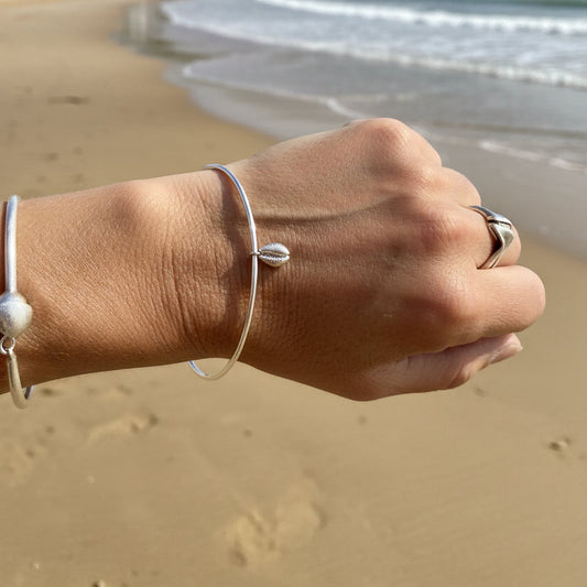A close-up of a person's wrist and hand wearing a silver bangle with a small cowry shell charm, against a plain light background.