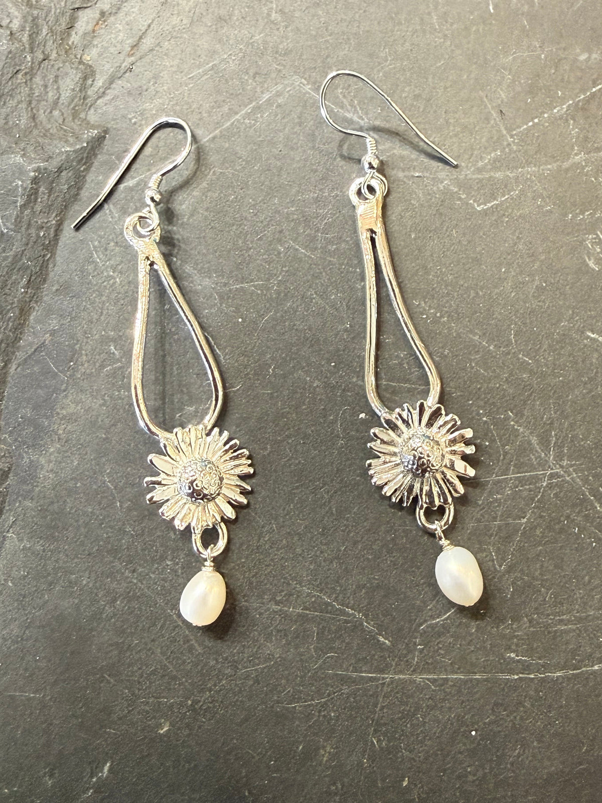 A pair of silver-toned dangle earrings with sunflower charms and white teardrop beads, displayed on a dark textured slate surface.
