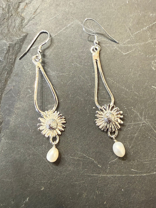A pair of silver-toned dangle earrings with sunflower charms and white teardrop beads, displayed on a dark textured slate surface.