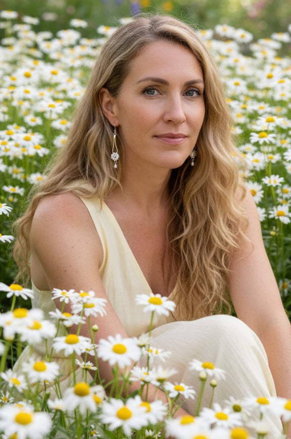 A close-up side profile of a woman wearing a long silver dangle earring with a white floral center and pearl drop, in a country garden