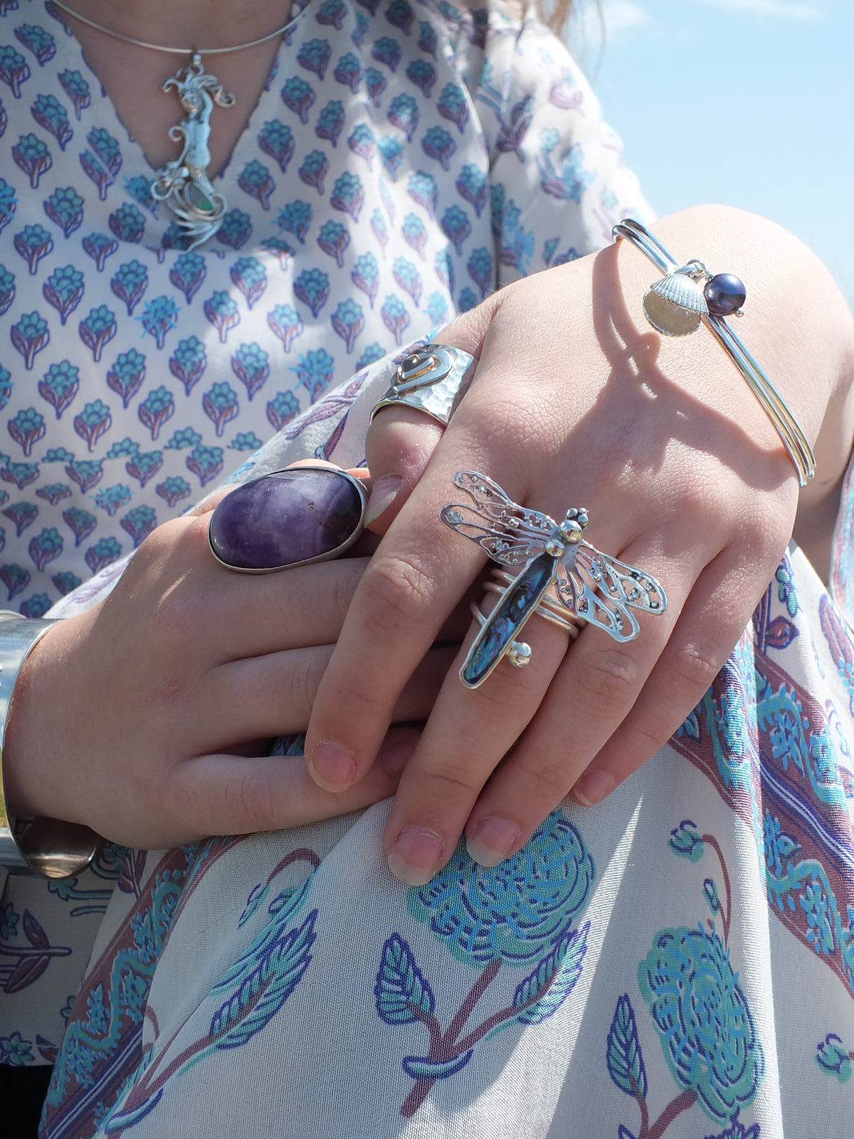 A close-up of a person's hands wearing multiple silver-toned rings and bracelets, including a dragonfly ring and a bangle with a blue bead, while wearing a white and blue patterned dress outdoors.