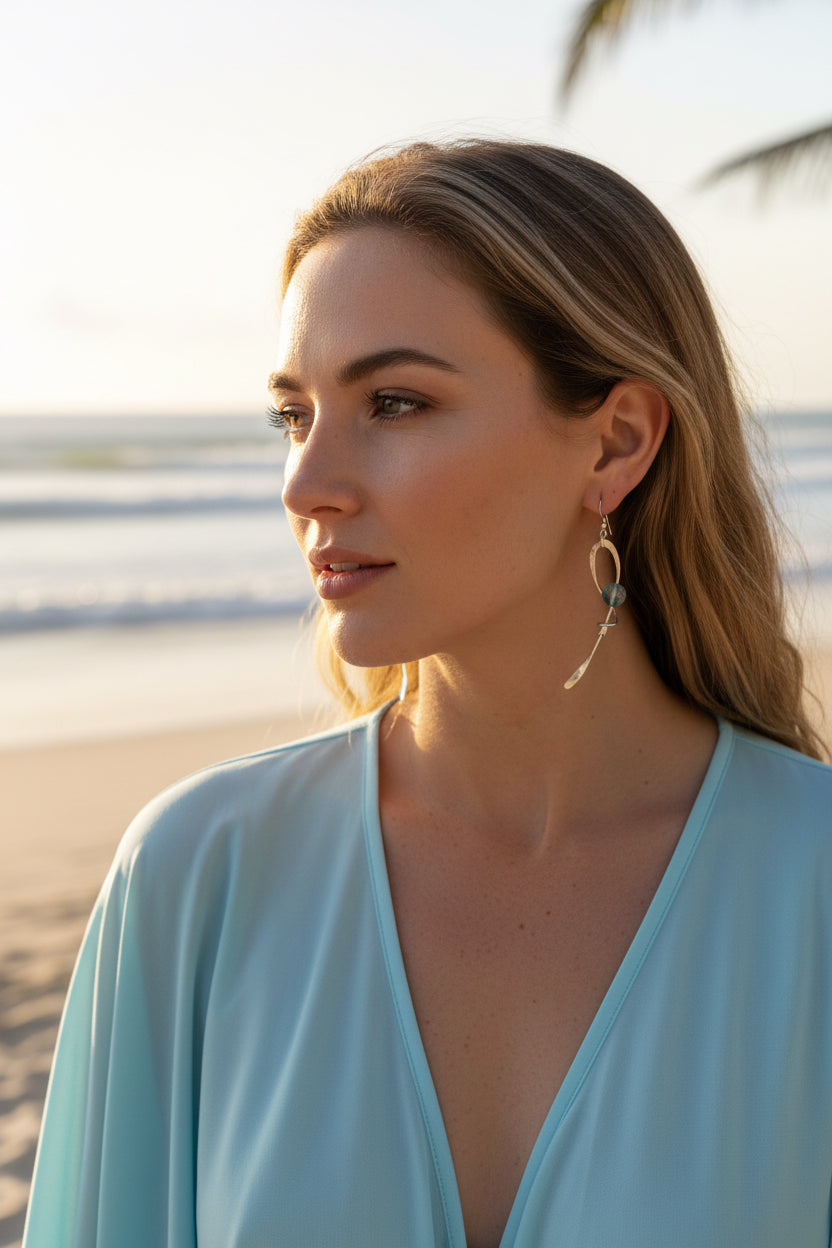 A close-up of a person's ear and neck wearing a long, sculptural silver dangle earring with a polished oval loop, a small clear stone, and a curved silver drop.
