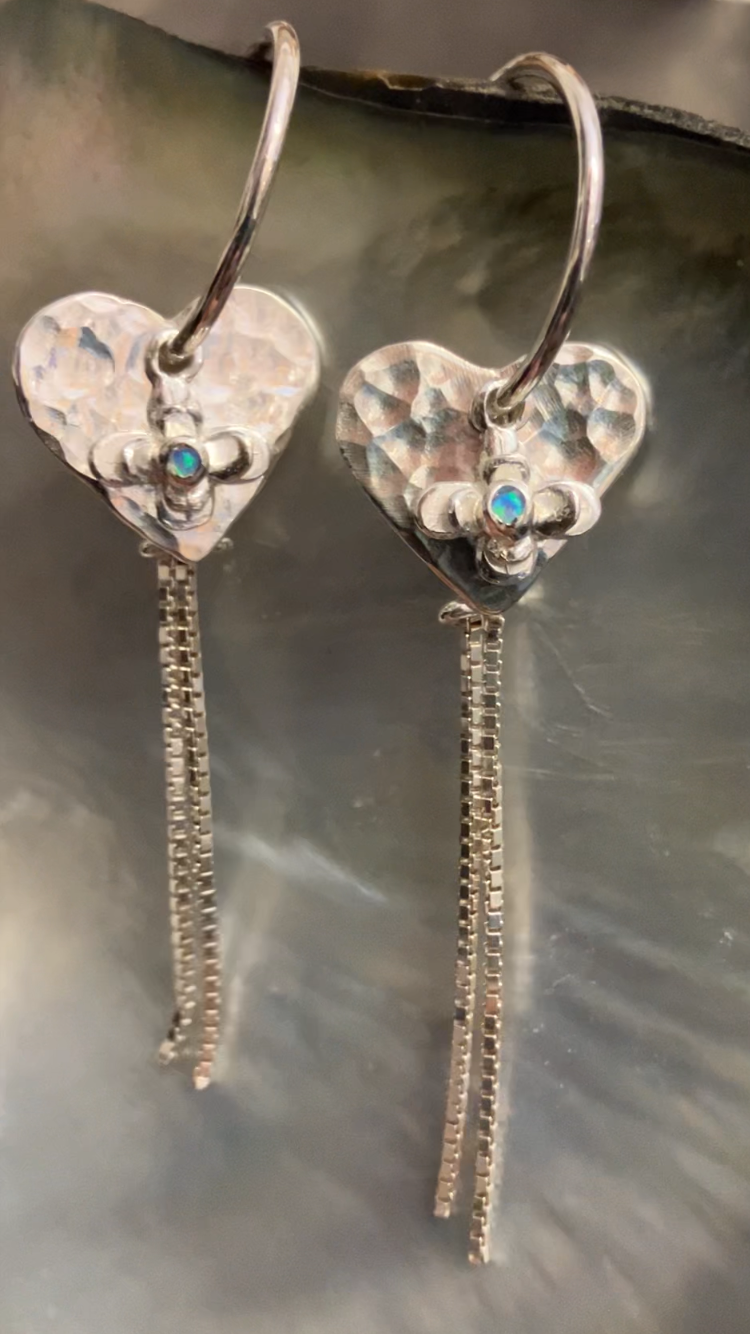 Heart-shaped earrings with dangling elements worn by a person on a beach at sunset.