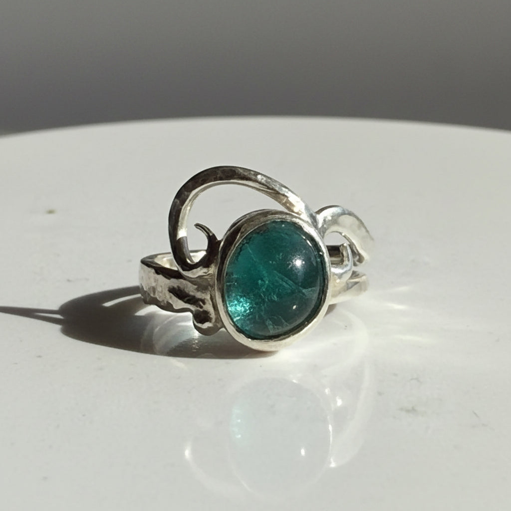 A silver ring with a textured organic band and a round green Seaglass gemstone, resting on a light reflective surface.